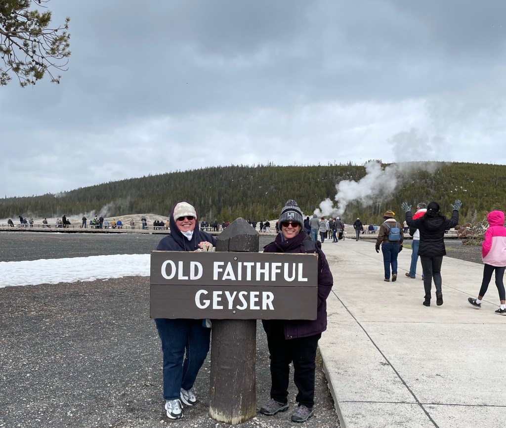 Us standing in front of sign reading “Old Faithful Geyser”