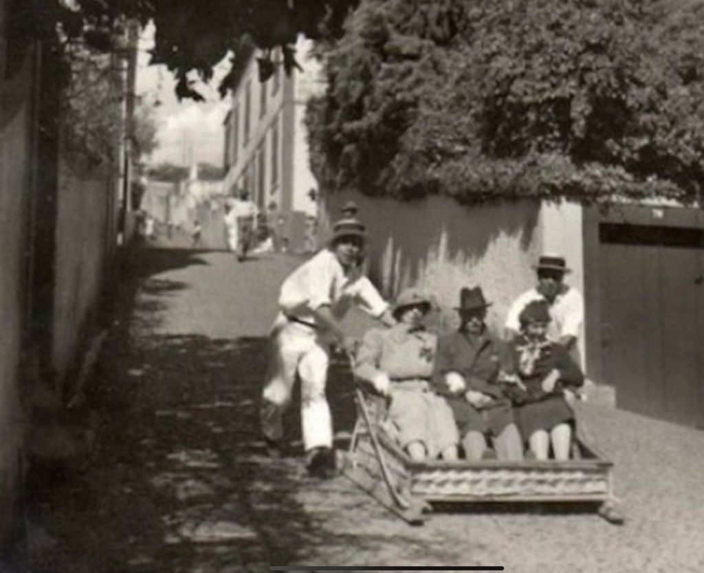 Three people being sledded down the street in a basket back in the day.  What day, I do not know.  But they’re wearing clothes that appear to be 1930s-ish.