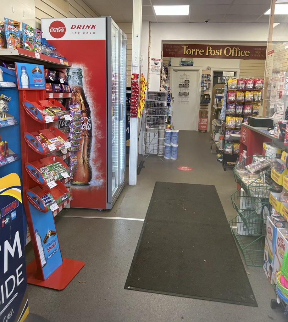 The inside of a convenience store with a post office counter at the back. The old sign reads “Established 10th June 1839 Torre Post Office Torquay’s first sub-office”